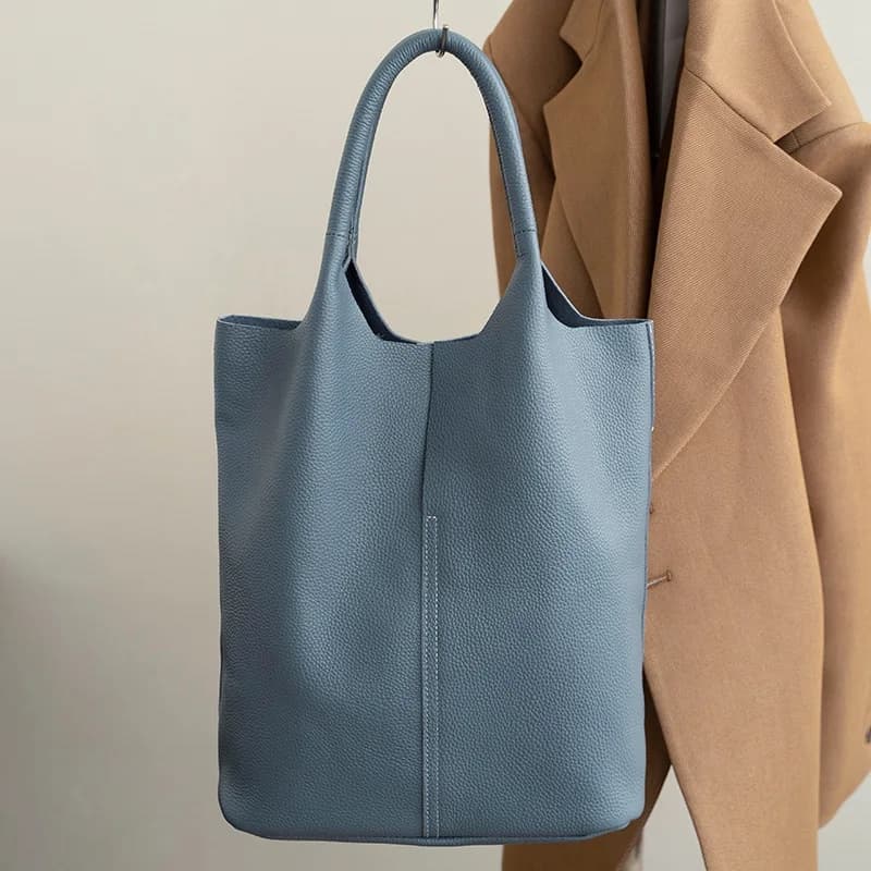 A blue soft leather tote bag with two rounded handles hanging by a coat